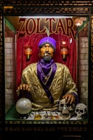 A fortune teller machine featuring a mannequin dressed in ornate clothing with a purple turban stands behind glass. The figure is adorned with jewelry and appears to hold a glowing crystal ball. The background is draped in red curtains, and there are decorative skulls on the table in front of the machine.