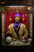 A fortune teller machine featuring a mannequin dressed in ornate clothing with a purple turban stands behind glass. The figure is adorned with jewelry and appears to hold a glowing crystal ball. The background is draped in red curtains, and there are decorative skulls on the table in front of the machine.