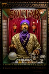 A fortune teller machine featuring a mannequin dressed in ornate clothing with a purple turban stands behind glass. The figure is adorned with jewelry and appears to hold a glowing crystal ball. The background is draped in red curtains, and there are decorative skulls on the table in front of the machine.