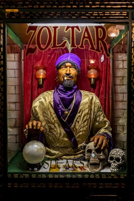 A fortune teller machine featuring a mannequin dressed in ornate clothing with a purple turban stands behind glass. The figure is adorned with jewelry and appears to hold a glowing crystal ball. The background is draped in red curtains, and there are decorative skulls on the table in front of the machine.