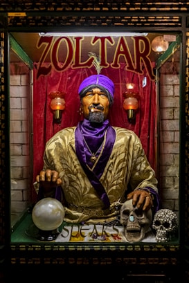 A fortune teller machine featuring a mannequin dressed in ornate clothing with a purple turban stands behind glass. The figure is adorned with jewelry and appears to hold a glowing crystal ball. The background is draped in red curtains, and there are decorative skulls on the table in front of the machine.