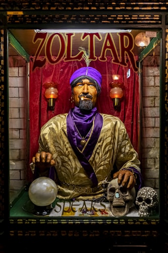 A fortune teller machine featuring a mannequin dressed in ornate clothing with a purple turban stands behind glass. The figure is adorned with jewelry and appears to hold a glowing crystal ball. The background is draped in red curtains, and there are decorative skulls on the table in front of the machine.