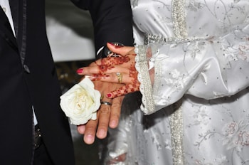 A close-up of a couple's hands adorned in intricate henna designs and jewelry, including rings, holding a white rose. The person on the left wears a dark suit while the other wears a white, embroidered and embellished garment.