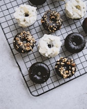 A collection of chocolate and vanilla donuts is arranged on a black cooling rack. Some donuts are topped with white icing and almond slices, while others have a chocolate glaze sprinkled with nuts. The background is a light gray surface.