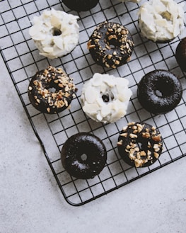 A collection of chocolate and vanilla donuts is arranged on a black cooling rack. Some donuts are topped with white icing and almond slices, while others have a chocolate glaze sprinkled with nuts. The background is a light gray surface.