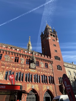 A grand red building with ornate architectural details and intricate murals covers the facade. A tall clock tower rises on the right, its red brick contrasting with the blue sky above. The structure is richly decorated with sculptures and carvings. Near the bottom, a red kiosk with a 'Piadina Stand' sign is visible, and there is a white vehicle parked nearby.