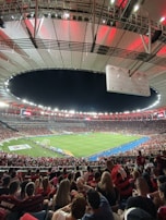 A vibrant and crowded soccer stadium filled with fans wearing red and black jerseys. The field is illuminated under bright stadium lights, and people are gathered closely, creating an energetic atmosphere. The roof structure is visible and casts a red glow over the crowd.