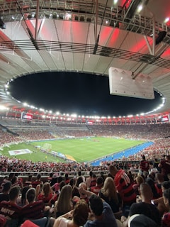 A vibrant and crowded soccer stadium filled with fans wearing red and black jerseys. The field is illuminated under bright stadium lights, and people are gathered closely, creating an energetic atmosphere. The roof structure is visible and casts a red glow over the crowd.