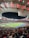 A vibrant and crowded soccer stadium filled with fans wearing red and black jerseys. The field is illuminated under bright stadium lights, and people are gathered closely, creating an energetic atmosphere. The roof structure is visible and casts a red glow over the crowd.