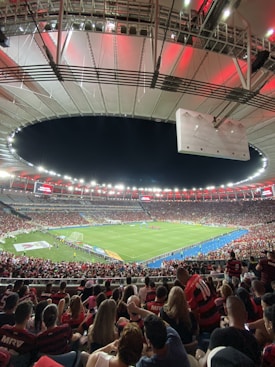 A vibrant and crowded soccer stadium filled with fans wearing red and black jerseys. The field is illuminated under bright stadium lights, and people are gathered closely, creating an energetic atmosphere. The roof structure is visible and casts a red glow over the crowd.