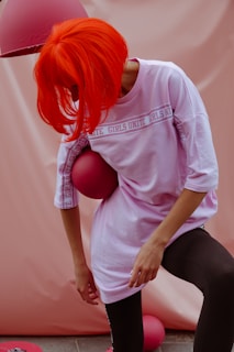 A person with vibrant orange hair leans forward, holding a pink ball against their side. They are wearing a light pink oversized shirt with the words 'Girls Unite' repeated on it, paired with black leggings. The background is a soft pink tone and there's a pink lampshade above.