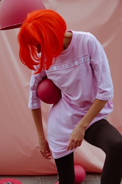 A person with vibrant orange hair leans forward, holding a pink ball against their side. They are wearing a light pink oversized shirt with the words 'Girls Unite' repeated on it, paired with black leggings. The background is a soft pink tone and there's a pink lampshade above.