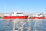 Two red and white pilot boats are docked at a harbor, with a clear blue sky above and a calm body of water below. The hulls display the word 'PILOT' in bold black letters. A metal railing is visible in the foreground, suggesting the perspective from another boat or dock.