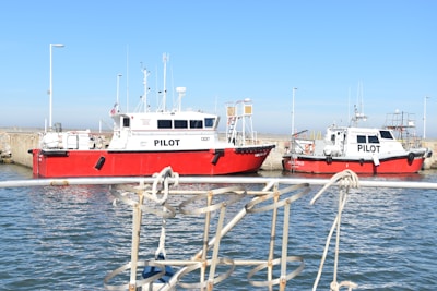 Two red and white pilot boats are docked at a harbor, with a clear blue sky above and a calm body of water below. The hulls display the word 'PILOT' in bold black letters. A metal railing is visible in the foreground, suggesting the perspective from another boat or dock.