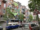 A vibrantly painted building with a colorful facade featuring various artworks, including abstract shapes, faces, and patterns. Several banners and signs with text hang from the balconies. A blue and white van is parked in front of the building, alongside other cars. Trees with green foliage line the street, and a traffic sign depicting construction is visible on the right.
