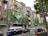 A vibrantly painted building with a colorful facade featuring various artworks, including abstract shapes, faces, and patterns. Several banners and signs with text hang from the balconies. A blue and white van is parked in front of the building, alongside other cars. Trees with green foliage line the street, and a traffic sign depicting construction is visible on the right.