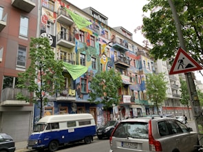 A vibrantly painted building with a colorful facade featuring various artworks, including abstract shapes, faces, and patterns. Several banners and signs with text hang from the balconies. A blue and white van is parked in front of the building, alongside other cars. Trees with green foliage line the street, and a traffic sign depicting construction is visible on the right.