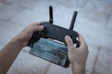 Hands are holding a drone controller with a smartphone attached below it, displaying a live aerial view of a city landscape. The setup appears to be used for flying a drone.