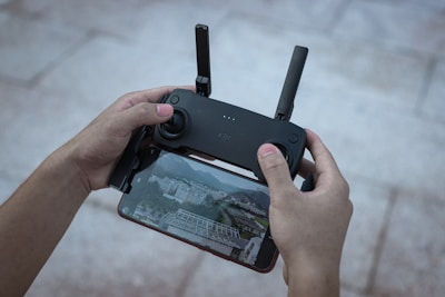 Hands are holding a drone controller with a smartphone attached below it, displaying a live aerial view of a city landscape. The setup appears to be used for flying a drone.