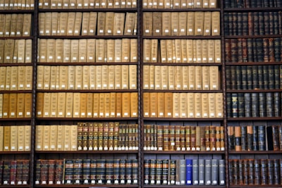 Shelves filled with books organized in a library. The books are arranged in a neat, orderly fashion, with several rows of similarly bound volumes. The upper shelves display books predominantly in beige or tan covers, while the lower shelves have a mix of colorful spines.