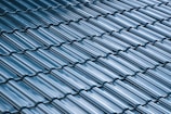 A close-up view of a roof with glossy, blue ceramic tiles arranged in a uniform, overlapping pattern. The tiles have a curved design and the surface reflects light, creating a shiny appearance.