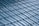 A close-up view of a roof with glossy, blue ceramic tiles arranged in a uniform, overlapping pattern. The tiles have a curved design and the surface reflects light, creating a shiny appearance.