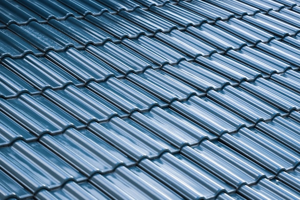 A close-up view of a roof with glossy, blue ceramic tiles arranged in a uniform, overlapping pattern. The tiles have a curved design and the surface reflects light, creating a shiny appearance.