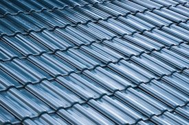 A close-up view of a roof with glossy, blue ceramic tiles arranged in a uniform, overlapping pattern. The tiles have a curved design and the surface reflects light, creating a shiny appearance.
