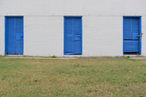 Three blue metal doors with horizontal slats are evenly spaced along a white brick wall. Each door is secured with a bolt and lock. The wall and doors form a linear pattern against the calm backdrop. In front of the wall, a strip of green grass with a few scattered leaves stretches across the foreground.