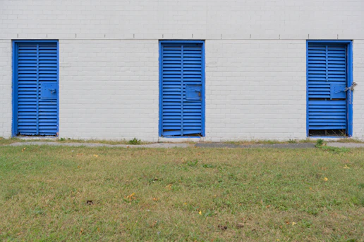 Three blue metal doors with horizontal slats are evenly spaced along a white brick wall. Each door is secured with a bolt and lock. The wall and doors form a linear pattern against the calm backdrop. In front of the wall, a strip of green grass with a few scattered leaves stretches across the foreground.