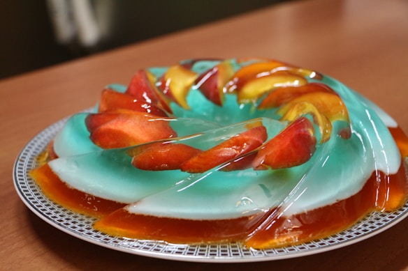 A beautifully arranged dessert featuring a translucent gelatin mold with slices of red and yellow fruit embedded within. The gelatin has a light blue hue and sits on a decorative plate with geometric patterns, placed on a wooden table.