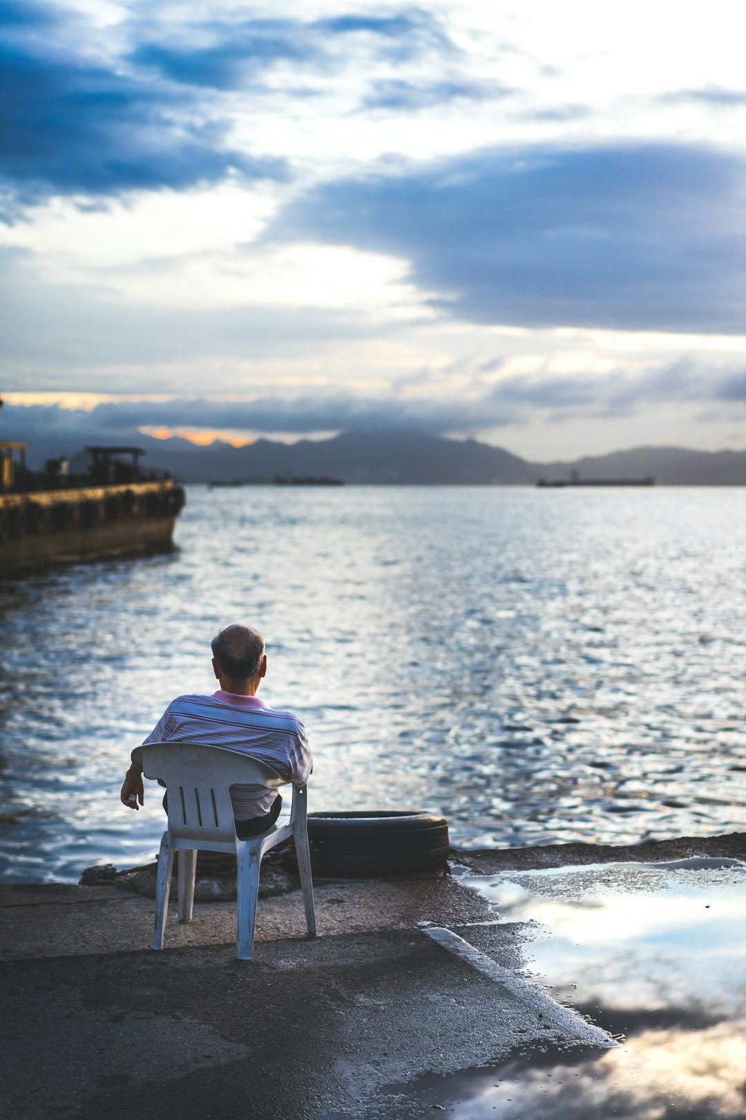 man in white shirt sitting on white plastic chair near body of water during daytime