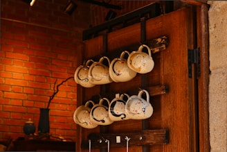A collection of white ceramic mugs with artistic designs hangs on a wooden rack against a brick wall. The mugs are arranged in two rows, and each has a unique design, featuring colorful paint splashes. The warm lighting and natural wood tones create a cozy atmosphere. A decorative vase and other small items are visible on a shelf in the background.