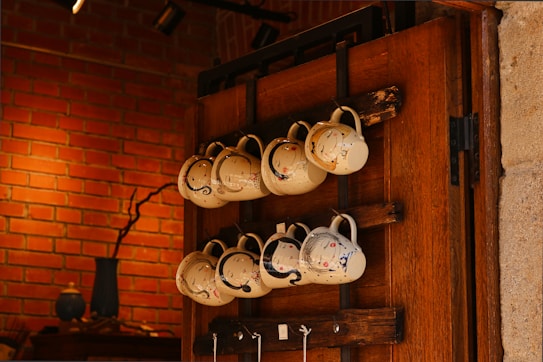 A collection of white ceramic mugs with artistic designs hangs on a wooden rack against a brick wall. The mugs are arranged in two rows, and each has a unique design, featuring colorful paint splashes. The warm lighting and natural wood tones create a cozy atmosphere. A decorative vase and other small items are visible on a shelf in the background.