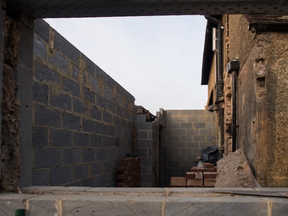 A partially constructed alleyway between two brick buildings, with stacks of bricks and construction materials scattered around. The walls are made of dark gray concrete blocks, and the right side has an older, textured stone surface.