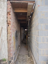 A narrow alleyway between two buildings is under construction, with exposed brick walls on one side and concrete blocks on the other. Metal scaffolding and wooden beams are used to support the structure, and construction tools and materials are visible along the path.