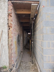 A narrow alleyway between two buildings is under construction, with exposed brick walls on one side and concrete blocks on the other. Metal scaffolding and wooden beams are used to support the structure, and construction tools and materials are visible along the path.