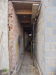 A narrow alleyway between two buildings is under construction, with exposed brick walls on one side and concrete blocks on the other. Metal scaffolding and wooden beams are used to support the structure, and construction tools and materials are visible along the path.