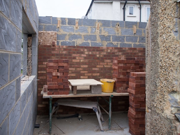 Construction site featuring an unfinished wall made of red and grey bricks. Several stacks of bricks are placed on either side of a wooden table. A yellow bucket sits on top of the table. Partially constructed walls and a window frame can be seen surrounding the area.