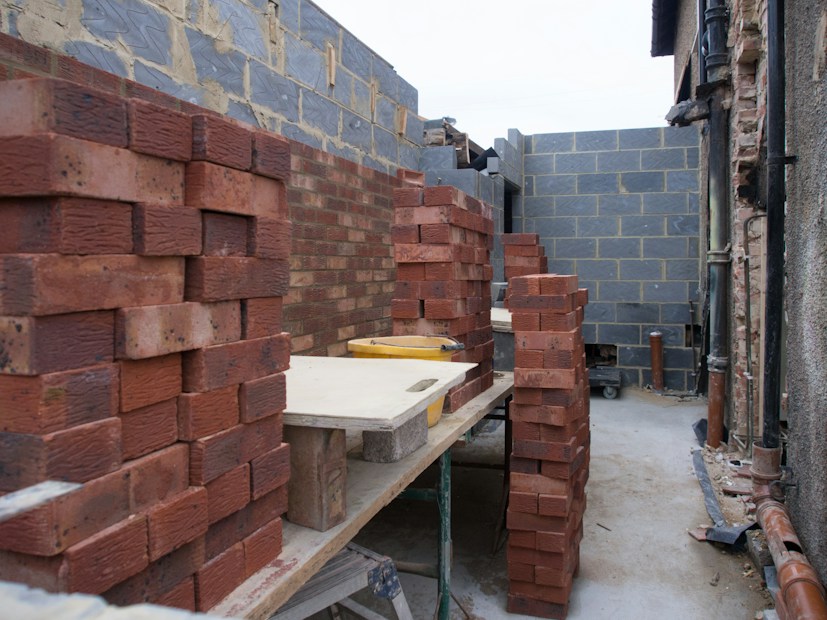 Stacks of red bricks are neatly arranged in a partially constructed building area. The walls are in various stages of construction, with some surfaces covered in unfinished plaster. There are tables holding bricks in preparation for further building work.