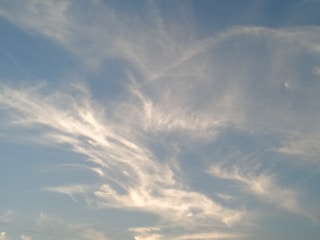 Light clouds are scattered across a clear blue sky. The clouds have a wispy texture and appear to be illuminated by the sunlight, giving a soft, serene appearance.