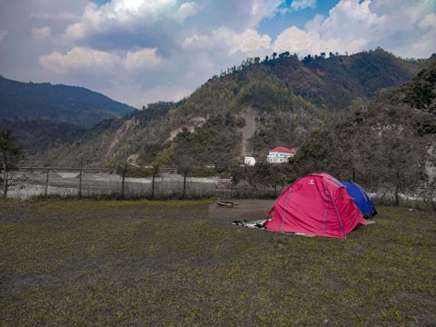 Several colorful tents are set up on a grassy area with mountains and a cloudy sky in the background. A building with a red roof and white walls is visible in the distance nestled among the forested hillside.