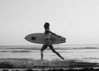 grayscale photo of woman holding surfboard on beach