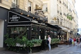 A Parisian café with ornate black awnings, an outdoor seating area covered with greenery, and a decorative gold snail sculpture on top. The setting includes classic Parisian architecture with cream-colored buildings and wrought-iron balconies. People are walking on the sidewalk and appear to be enjoying the bustling atmosphere.