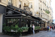 A Parisian café with ornate black awnings, an outdoor seating area covered with greenery, and a decorative gold snail sculpture on top. The setting includes classic Parisian architecture with cream-colored buildings and wrought-iron balconies. People are walking on the sidewalk and appear to be enjoying the bustling atmosphere.
