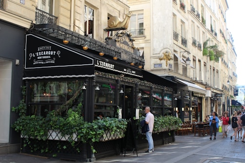 A Parisian café with ornate black awnings, an outdoor seating area covered with greenery, and a decorative gold snail sculpture on top. The setting includes classic Parisian architecture with cream-colored buildings and wrought-iron balconies. People are walking on the sidewalk and appear to be enjoying the bustling atmosphere.