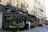 A Parisian café with ornate black awnings, an outdoor seating area covered with greenery, and a decorative gold snail sculpture on top. The setting includes classic Parisian architecture with cream-colored buildings and wrought-iron balconies. People are walking on the sidewalk and appear to be enjoying the bustling atmosphere.