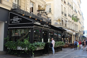 A Parisian café with ornate black awnings, an outdoor seating area covered with greenery, and a decorative gold snail sculpture on top. The setting includes classic Parisian architecture with cream-colored buildings and wrought-iron balconies. People are walking on the sidewalk and appear to be enjoying the bustling atmosphere.