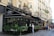 A Parisian café with ornate black awnings, an outdoor seating area covered with greenery, and a decorative gold snail sculpture on top. The setting includes classic Parisian architecture with cream-colored buildings and wrought-iron balconies. People are walking on the sidewalk and appear to be enjoying the bustling atmosphere.