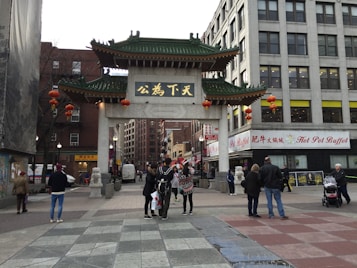 A traditional Chinese gate with ornate green roofing stands prominently in a bustling urban setting. Red lanterns are hung from the gate, which is inscribed with Chinese characters. People are gathered around, walking and taking photos. Surrounding the gate are modern buildings, one of which houses a 'Hot Pot Buffet.' The scene suggests a blend of cultural and modern urban elements.